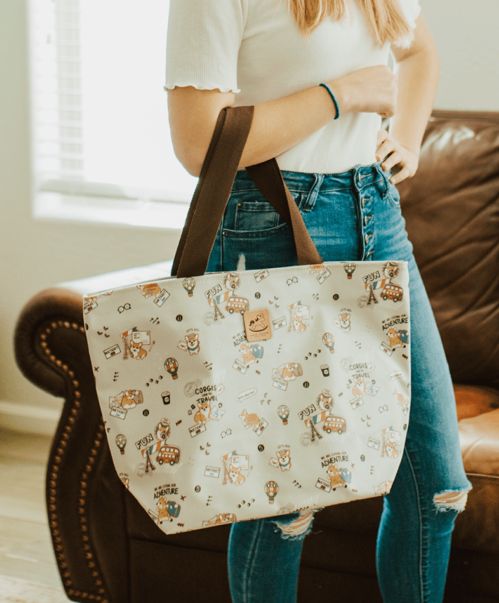 Girl wearing blue jeans and white shirt is carrying a gray corgi themed tote on her arm 